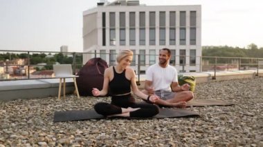 Genç mutlu çift yoga dersinden sonra modern evin çatısında dans ediyor. Atletik adam ve zayıf kadın sabah ısınması sırasında bacak bacak üstüne atarak oturuyorlar..
