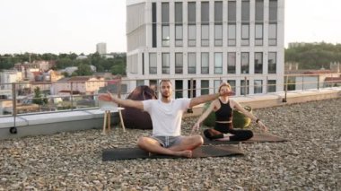 Fiziksel olarak güçlü erkek ve kadın minderlerin üzerinde oturuyor ve elleri yukarıda yoga yapıyorlar. Mutlu ve sağlıklı bir çift sabahları çatıda meditasyon yapıyor..