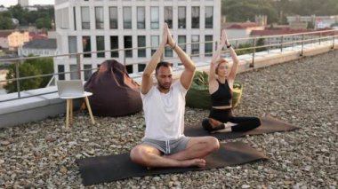 Sabahları çatıda meditasyon yapan sağlıklı bir çift. Fiziksel olarak güçlü erkek ve kadın minderlerin üzerinde oturuyor ve elleri yukarıda yoga yapıyorlar.. .