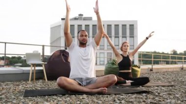 Sabahları çatıda meditasyon yapan sağlıklı bir çift. Fiziksel olarak güçlü erkek ve kadın minderlerin üzerinde oturuyor ve elleri yukarıda yoga yapıyorlar.. .