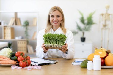 Çalışma masasında otururken beyaz önlüklü, elinde mikro yeşillik kabıyla çekici bir bayanın yakın görüntüsü. Kafkasyalı beslenme uzmanı beslenme düzenine modern ekleme öneriyor.