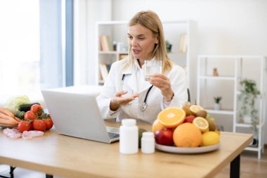 Deneyimli kadın beslenme uzmanı, bir online oturum sırasında hastaya dizüstü bilgisayar kullanarak suyun faydalarından bahsediyor. Beyaz önlüklü kadın doktor bir video görüşmesi sırasında elinde bir bardak su tutuyordu..