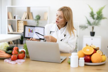 Grafikler yoluyla beslenme kılavuzu sağlamada etkili bir yetişkin uzmanı. Doktor önlüklü kendine güvenen bir kadın klinikte mesai saatleri boyunca laptoptan konferans görüşmesiyle sunum yapıyor..