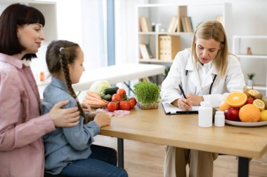 Deneyimli kadın beslenme düzeni, küçük çocuklar için sağlıklı beslenme kursuna hazırlanıyor. Olgun beyaz kadın doktor, çocuk kızıyla konuşuyor dizlerinin üstüne çöküp diyet planı yazıyor..