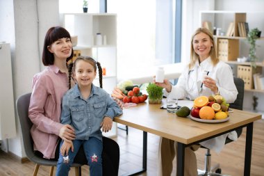 Kadın beslenme uzmanının portresi elinde haplarla annesinin kucağında oturan ve kameraya bakan küçük kız için besin takviyesi yapıyor. Olgun çocuk doktoruna modern klinikte kadın hasta kabul edilir.