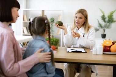 Elinde taze avokadosu olan olgun bir kadın. Küçük kız çocuğu ve annesiyle danışma odasında sohbet ediyor. Besin bolluğuyla beslenme düzenini geliştirmek için tavsiye veren deneyimli tıp uzmanı.