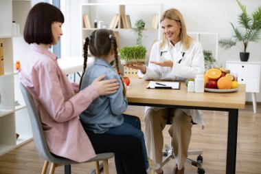 Yetişkin kadın beslenme uzmanıyla randevuda annesinin kucağında oturan küçük kız sağlıklı beslenme planı yapıyor. Hasta diyetine eklerken mikroyeşili tutan güzel kadın doktor..