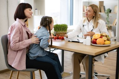 Hasta diyetine eklerken mikroyeşili tutan güzel bir kadın doktor. Yetişkin kadın beslenme uzmanıyla randevuda annesinin kucağında oturan küçük kız sağlıklı beslenme planı yapıyor..