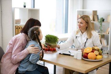 Hasta diyetine eklerken mikroyeşili tutan güzel bir kadın doktor. Yetişkin kadın beslenme uzmanıyla randevuda annesinin kucağında oturan küçük kız sağlıklı beslenme planı yapıyor..