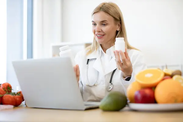 Caucasian doctor distantly describes the treatment plan. Positive mature nutritionist woman holding pills or vitamins in hands during online consultation with patient.