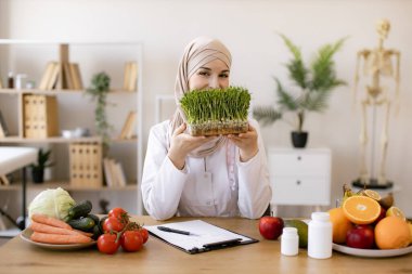 Çalışma masasında otururken beyaz önlüklü, elinde mikro yeşillik kabıyla çekici bir bayanın yakın görüntüsü. Müslüman beslenme uzmanı tesettüre modern ekleme öneriyor.