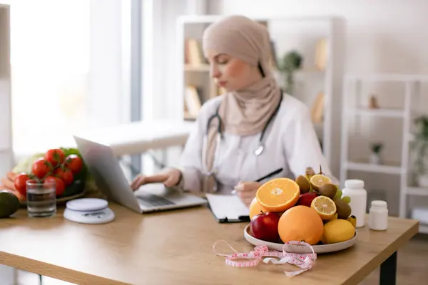 Serious Muslim lady in doctors coat typing on modern laptop in consulting room of medical center. Experienced nutrition professional searching for weight loss information on food websites.