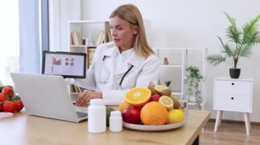 Doktor önlüklü kendine güvenen bayan klinikte mesai saatleri boyunca laptoptan konferans görüşmesiyle sunum yapıyor. Grafikler aracılığıyla beslenme kılavuzu sağlamada etkili bir yetişkin uzmanı..