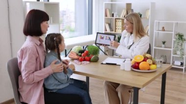 Anasının kucağında oturan küçük liseli kız, kadın pediatrist beslenme uzmanıyla görüşüyor. Olgun beyaz kadın doktor sağlıklı beslenmek için tablet grafiklerinde görünüyor..