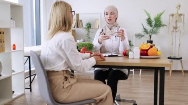 Müslüman kadın doktor, beslenme sıvılarını yenilemenin önemini göstermek için bir bardak su tutuyor. Olgun kadın hasta, beslenme uzmanını ziyaret ederken su dengesi hakkında bilgi alır..