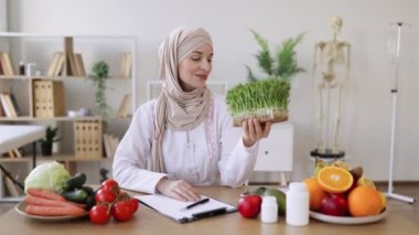 Çalışma masasında otururken beyaz önlüklü, elinde mikro yeşillik kabıyla çekici bir bayanın yakın görüntüsü. Müslüman beslenme uzmanı tesettüre modern ekleme öneriyor.