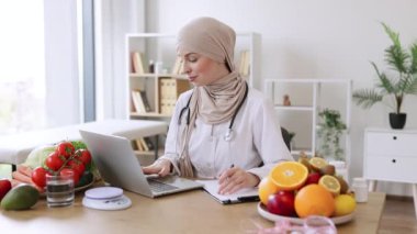 Gıda sitelerinde kilo verme bilgisi arayan deneyimli bir beslenme uzmanı. Tıp merkezinin danışma odasında modern dizüstü bilgisayarda yazan doktor ceketli ciddi bir Müslüman kadın..