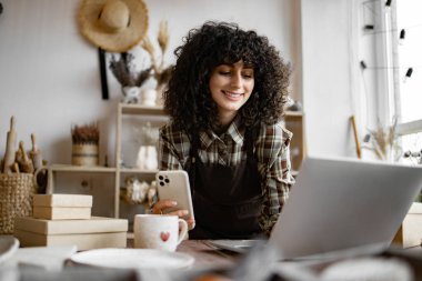 Kıvırcık genç kadın çömlekçi, kendi çömlekçisinin web sitesi için kilden el yapımı kupaların fotoğraflarını çekiyor. Beyaz kadın web sayfası ve sosyal medya portföyünü güncelledi.