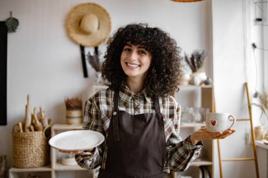 Profesyonel satıcı el ele tutuşan bardak ve tabakta hasar olup olmadığını kontrol ediyor. Kahverengi önlük giyen çekici kıvırcık kadın modern dekor mağazasından mal alıyor..