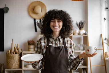 Profesyonel satıcı el ele tutuşan bardak ve tabakta hasar olup olmadığını kontrol ediyor. Kahverengi önlük giyen çekici kıvırcık kadın modern dekor mağazasından mal alıyor..