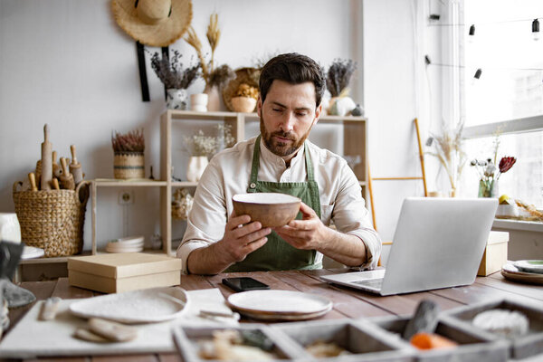 Взрослый владелец керамического магазина, используя ноутбук во время работы в магазине. Мужчина предприниматель, управляющий онлайн заказ кружку на сайте. Уверенный молодой бизнесмен, управляющий творческим малым бизнесом.