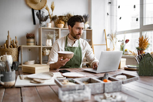 Взрослый владелец керамического магазина, используя ноутбук во время работы в магазине. Мужчина предприниматель, управляющий онлайн заказ кружку на сайте. Уверенный молодой бизнесмен, управляющий творческим малым бизнесом.
