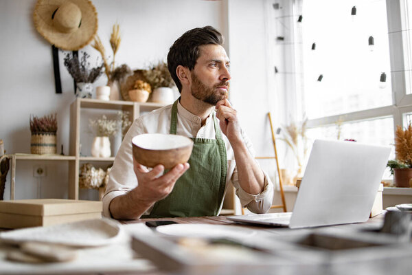 Молодой кавказский мужчина гончар доволен онлайн продажей чаши в Интернете на фоне полок с керамикой. Бородатый взрослый бизнесмен, занимающийся розничной торговлей посудой ручной работы.