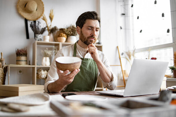 Молодой кавказский мужчина гончар доволен онлайн продажей чаши в Интернете на фоне полок с керамикой. Бородатый взрослый бизнесмен, занимающийся розничной торговлей посудой ручной работы.