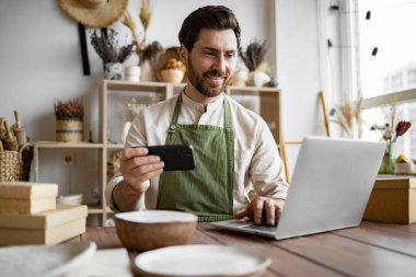 Dekoratif kil mağazasının pozitif yöneticisi telefonda sosyal medya için fotoğraf çekiyor. Olgun adam, çömlekçi, ev yapımı yemek yapmakla ve web sitesi portföyünü güncellemekle meşgul..