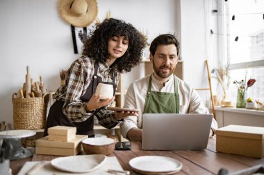 Kafkasyalı birkaç iş arkadaşı küçük çömlek imalatı işinde çalışıyor. Olgun erkek ve önlüklü genç kıvırcık saçlı kadın, mağaza sitesindeki ürünlerin envanterini çıkarmak için dizüstü bilgisayar kullanıyorlar..