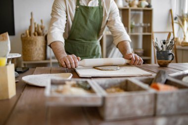 Parlak stüdyoda seramik sanatı yapma süreci ürünlerle dolu rafların arka planına karşı. Olgun sakallı erkek çömlekçi el yapımı yemekler yapmak için balçığı merdaneyle düzleştirir..
