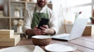 Dekoratif kil mağazasının pozitif yöneticisi telefonda sosyal medya için fotoğraf çekiyor. Olgun adam, çömlekçi, ev yapımı yemek yapmakla ve web sitesi portföyünü güncellemekle meşgul..
