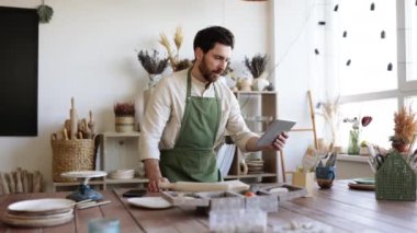 Önlüklü ve beyaz gömlekli sakallı yetişkin bir adam çömlekçilik ve seramik yemekler üzerine online kurs izliyor. Küçük işletme, el yapımı ürünler ve İnternet 'te online satışlar kavramı.