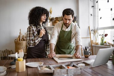 Yanındaki kadın el ele tutuşup ona çömlek atölyesinde ders verirken, adam elinde oklavayla kilden yuvarlanıyor. Stüdyo çeşitli çanak çömlek aletleri ve malzemelerle dolu, yaratıcı atmosfer yaratıyor..
