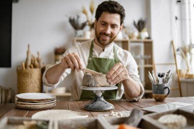 Sıcak ev çömlek atölyesinde kille çalışan gülen adam. Sakallı erkek, ahşap masaya oturmuş. Elinde çeşitli aletler, malzemeler ve dizüstü bilgisayarla iyi dekore edilmiş bir odada..