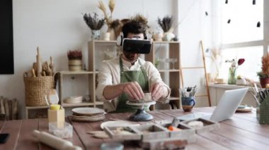 Sanatçı sanal gerçeklik gözlüğü takarken yaratıcı stüdyoda kilden vazo yontuyor. Çalışma alanı çeşitli araçlar, çanak çömlek ve sanat malzemeleriyle dolu, teknolojinin renklendirilmesi.