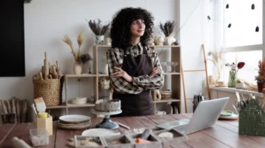 Önlüklü kendine güvenen kadın esnaf çömlek atölyesinde kollarını kavuşturmuş duruyor. Ahşap masa üzerinde çömlek aletleri ve dizüstü bilgisayarı olan yaratıcı çalışma alanı.