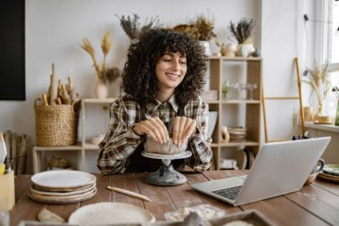 Dişi çömlekçi dizüstü bilgisayardan online ders izlerken stüdyoda seramik bir eser üzerinde çalışıyor. Kil, aletler ve stüdyo ekipmanlarıyla yaratıcı çalışma alanı.