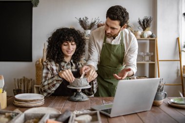Erkek, seramik atölyesinde kadınlara çömlek teknikleri öğretiyor. Kadın, erkeğin ona öğrettiği gibi kil parçasına konsantre oluyor. Pratik öğrenme ile yaratıcı hobi.