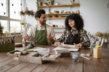 Sakallı olgun bir adam vazo heykeli yaparken kadınlar çömlek atölyesinde merdaneyle kil yuvarlıyor..