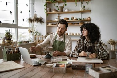 Önlüklü sakallı adam kadınlara çömlekçilik tekniklerini gösterirken yaratıcı atölyede çevrimiçi video dersi kullanarak kil yontuyor..