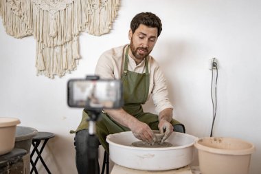 Genç yetişkin bir adam akıllı telefonuyla video blogu kaydederken seramik yapmak için çömlekçi tekerleği kullanıyor. Sanat ve zanaat sürecini bir ev stüdyosunda yakalamak..
