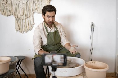 Olgun adam tekerlekli çömlek yapıyor ve video blogu kaydediyor. Çalışan çömlekçi stüdyoda zanaat gösterisi yapıyor..