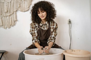 Young adult female potter using a pottery wheel to sculpt ceramic dishes in an art studio environment. clipart