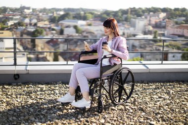 Tekerlekli sandalyedeki kendine güvenen kadın güneşli çatıda akıllı telefon kullanırken bir fincan kahve tutuyor. Bağımsızlığı, şehir yaşam tarzını ve modern teknolojiyi yakalıyor..