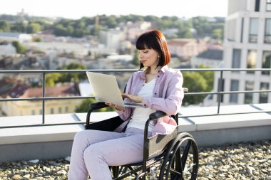 İş kadını, çevrimiçi bir çatı katında çalışırken manzaranın tadını çıkarıyor. Tekerlekli sandalyedeki olumlu beyaz kadın dizüstü bilgisayarla oturup uzaktan mesaj yazıyor..