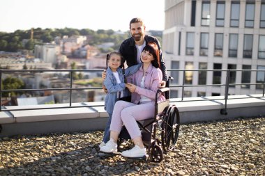 Özürlü genç bir ailenin portresi modern evlerle şehir planına karşı. Genç bir adam ve küçük bir kız tekerlekli sandalyede tedavi gören anneyle ilgileniyorlar..