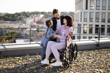 Tekerlekli sandalyedeki genç kadın modern evin terasında ailesiyle eğleniyor. Baba ve kızı, anneye ciddi yaralanma tedavisinde destek olurken boş zamanlarını temiz havada geçiriyorlar..