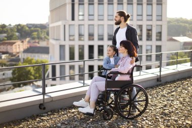 Sakallı beyaz bir baba, küçük tatlı bir kız ve genç bir anne modern evin çatısında günbatımını seyrediyor. Boş zaman, destek ve maluliyet kavramı.