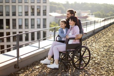 Boş zaman, destek ve maluliyet kavramı. Sakallı beyaz baba, küçük tatlı kız ve tekerlekli sandalyede genç anne modern evin çatısında gün batımını izliyorlar..